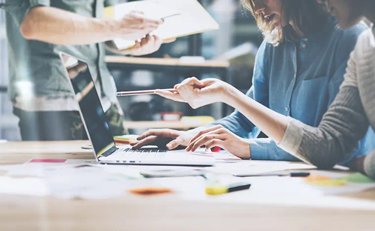 Two business women discuss something at their desk on a laptop.