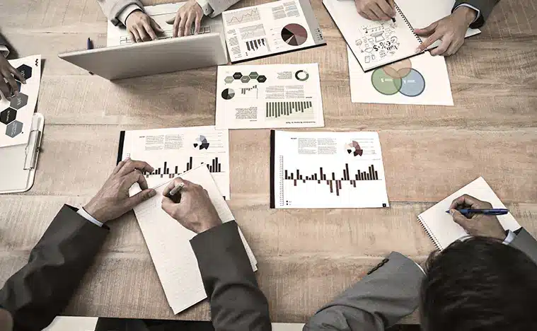 Reports and charts spread along a large conference table while they are being reviewed by several business people.